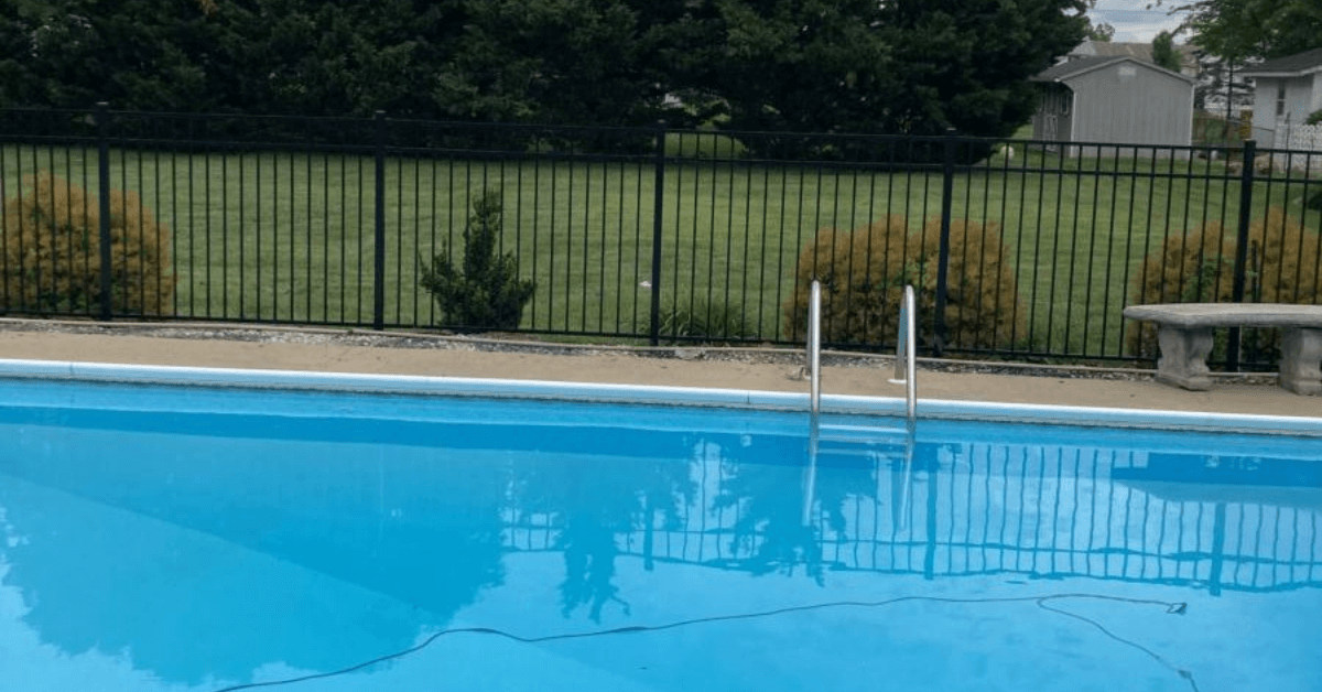 An empty outdoor swimming pool with bright turquoise water, metal fence enclosure, and diving board on a clear day.