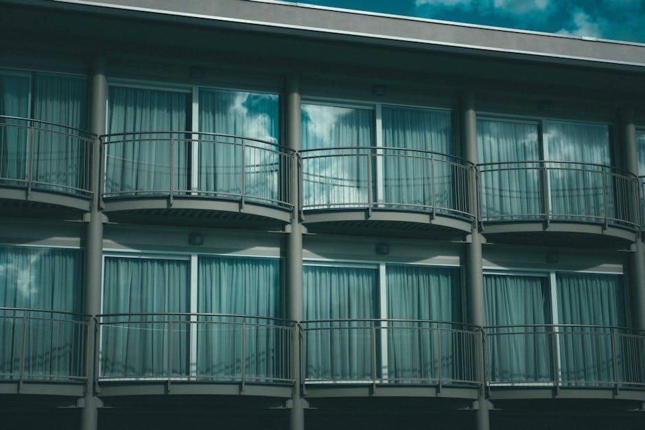 A modern apartment building facade with repeating rows of balconies and windows in teal-blue tones creating a geometric, minimalist architectural pattern.