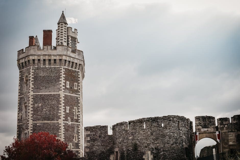 A historic medieval stone tower with crenellations rises against an overcast sky, with weathered castle walls and autumn ivy visible in the foreground.