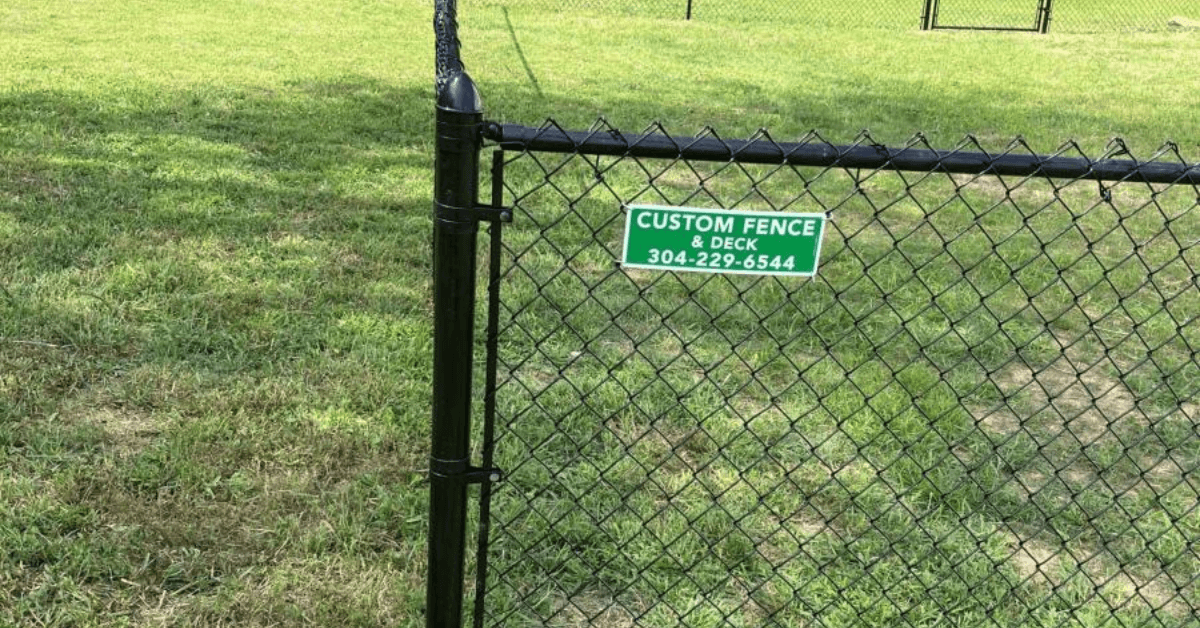 A close-up view of a black chain-link fence with a green directional sign attached, overlooking a grass field or sports facility.