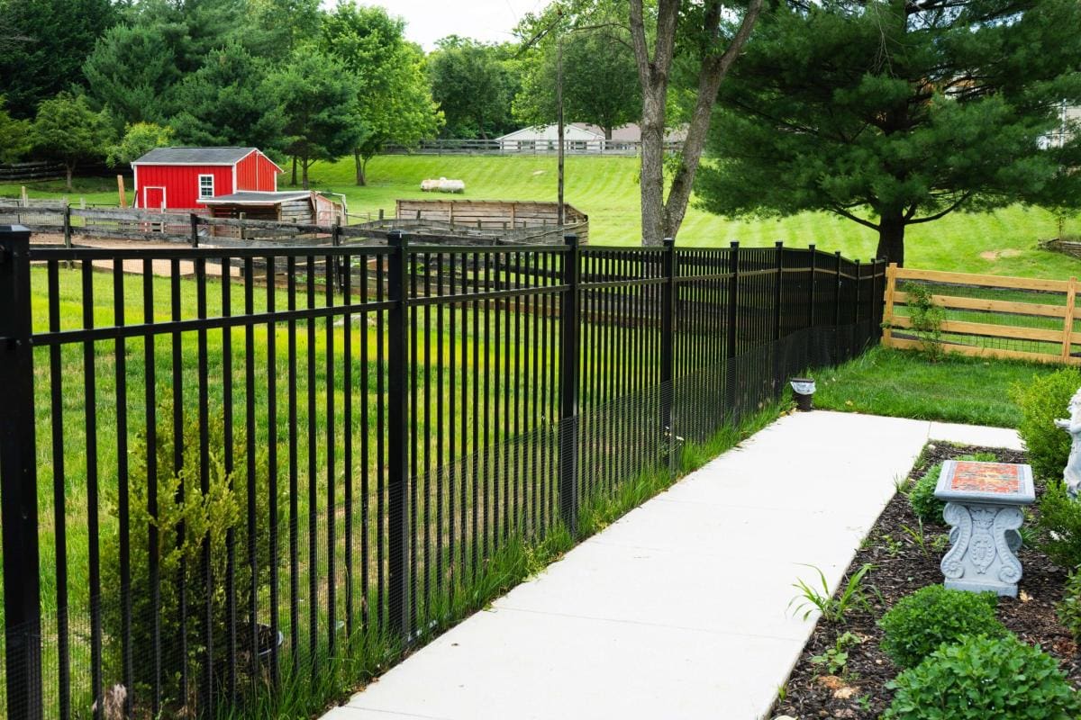 A peaceful rural property featuring a modern black metal fence alongside a white concrete pathway, with a red barn and manicured landscaping visible in a pastoral setting.