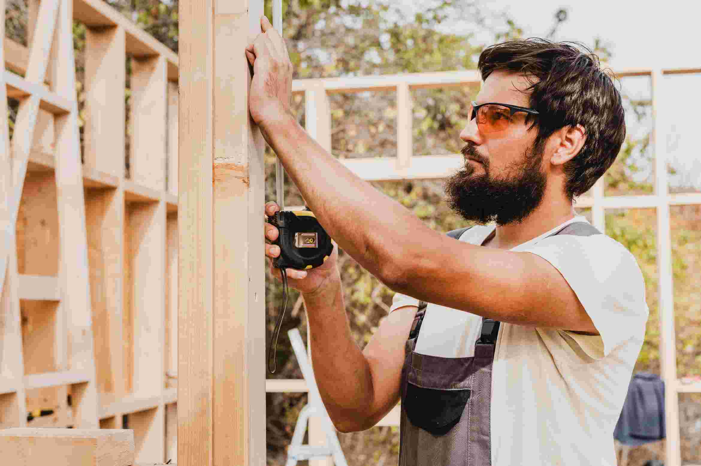 A bearded craftsman in a white tank top installs wooden cabinetry in a sunlit workshop using a power drill.