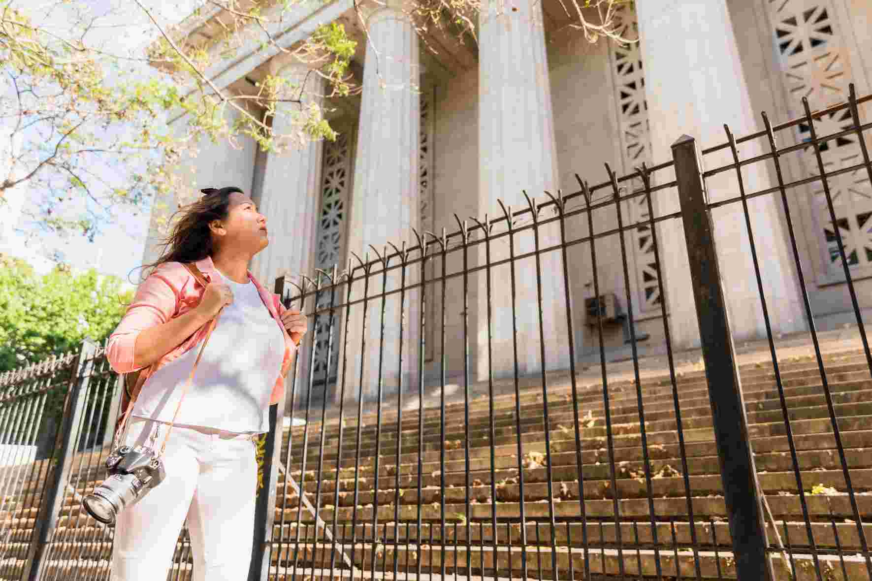 A woman in casual white attire stands at an ornate black iron gate with classical columns in the background during golden hour lighting.