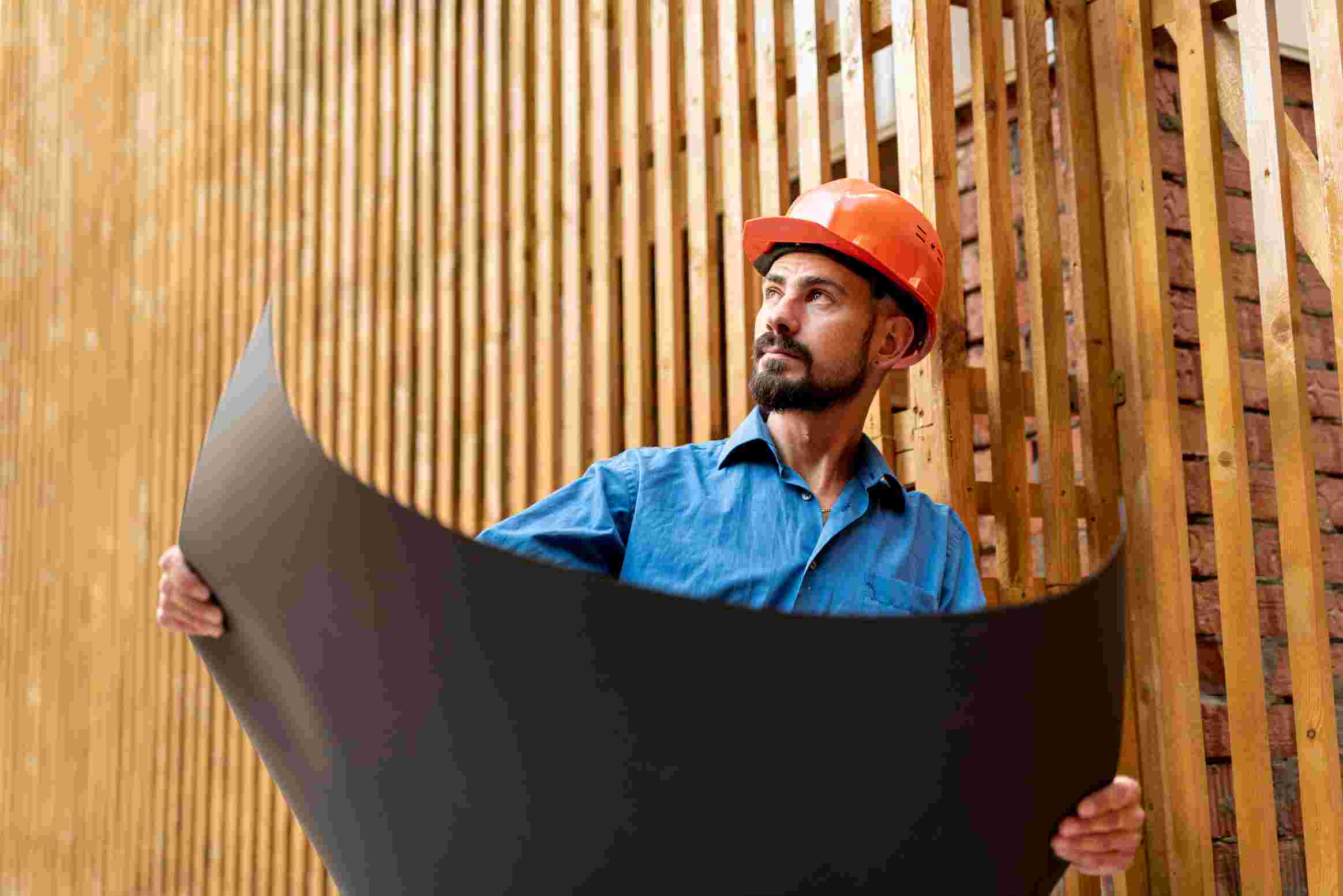 A construction worker wearing a red hard hat and blue shirt examines architectural blueprints against a wooden backdrop.