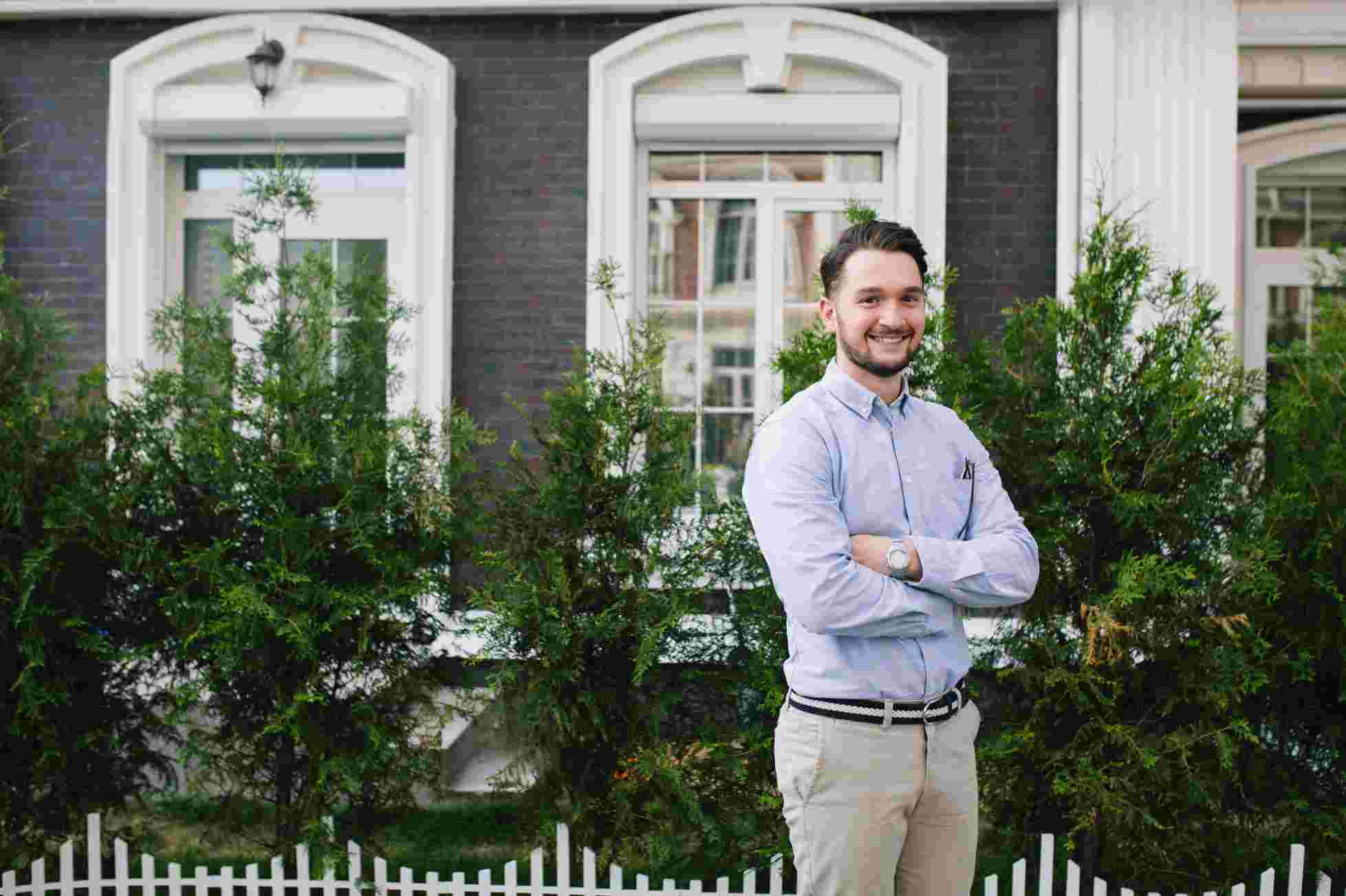 A man in a light blue shirt and khaki pants stands confidently in front of a residential building with white doors and manicured green hedges.
