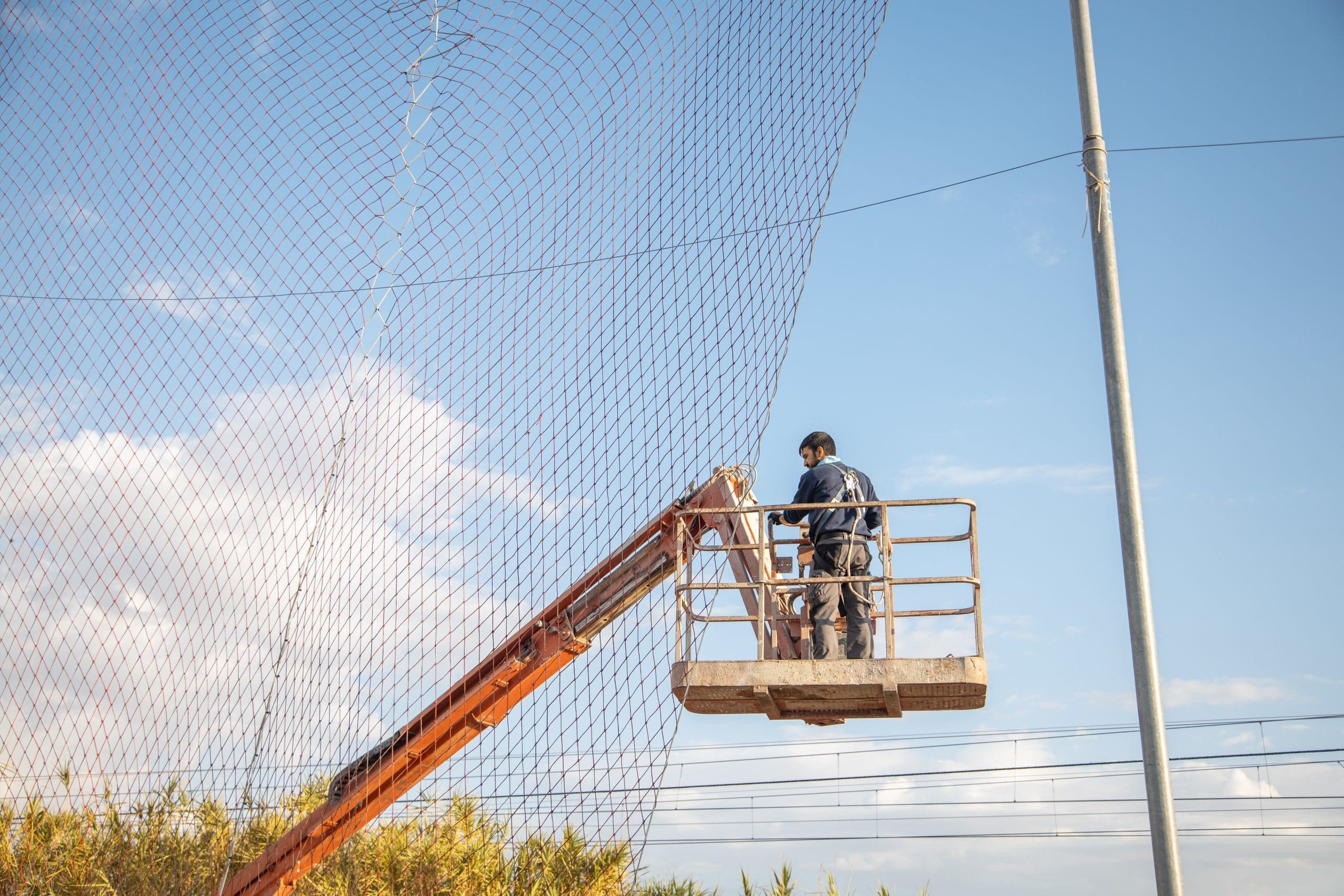 A worker in a yellow boom lift operates near a large safety net strung between utility poles against a partly cloudy sky.